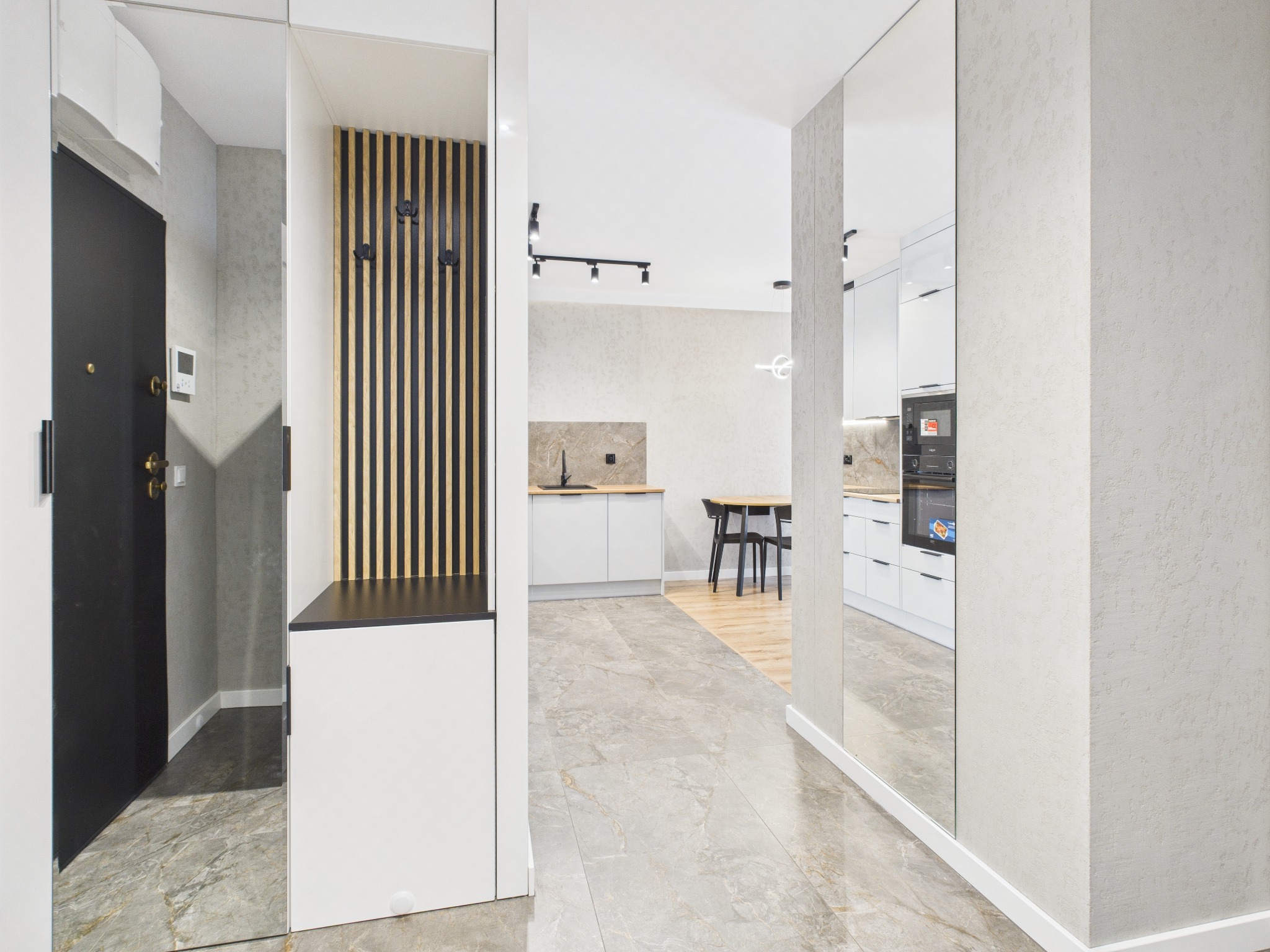 Nowoczesny przedpokój z lustrem, drewnianym panelem ściennym z wieszakami i marmurową podłogą w odcieniach szarości, z widokiem na białą kuchnię w tle.