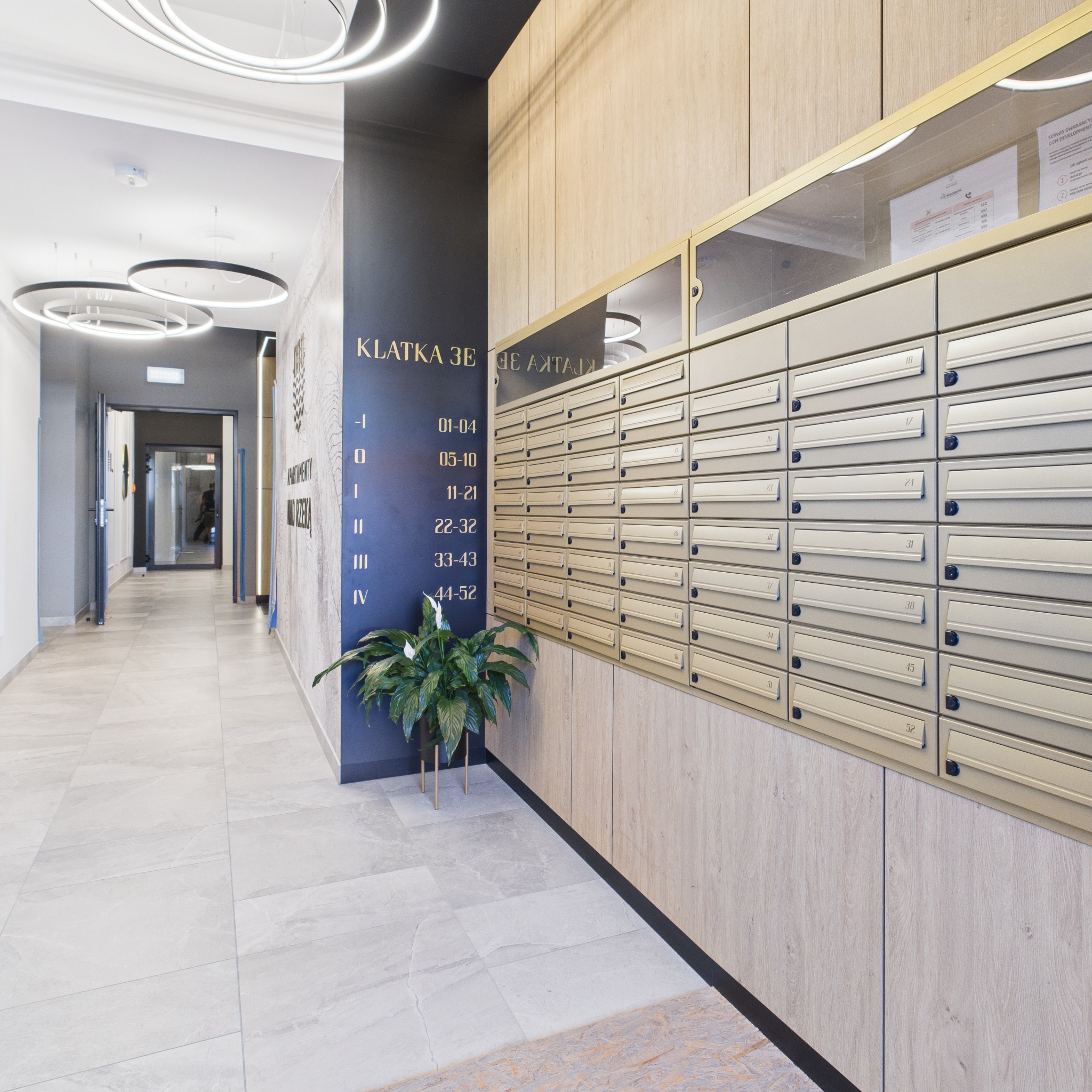 Nowoczesny hol apartamentowy z eleganckimi skrzynkami pocztowymi, numeracją mieszkań na ścianie i rośliną doniczkową. Oświetlenie sufitowe w kształcie okręgów.