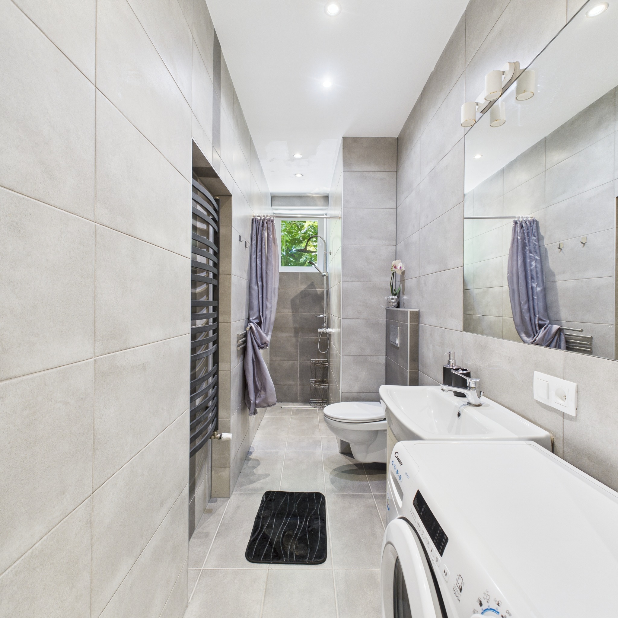 Jasna łazienka z szarymi płytkami, pralką Candy i grzejnikiem drabinkowym. Widoczna kabina prysznicowa z zasłoną i czarny dywanik.