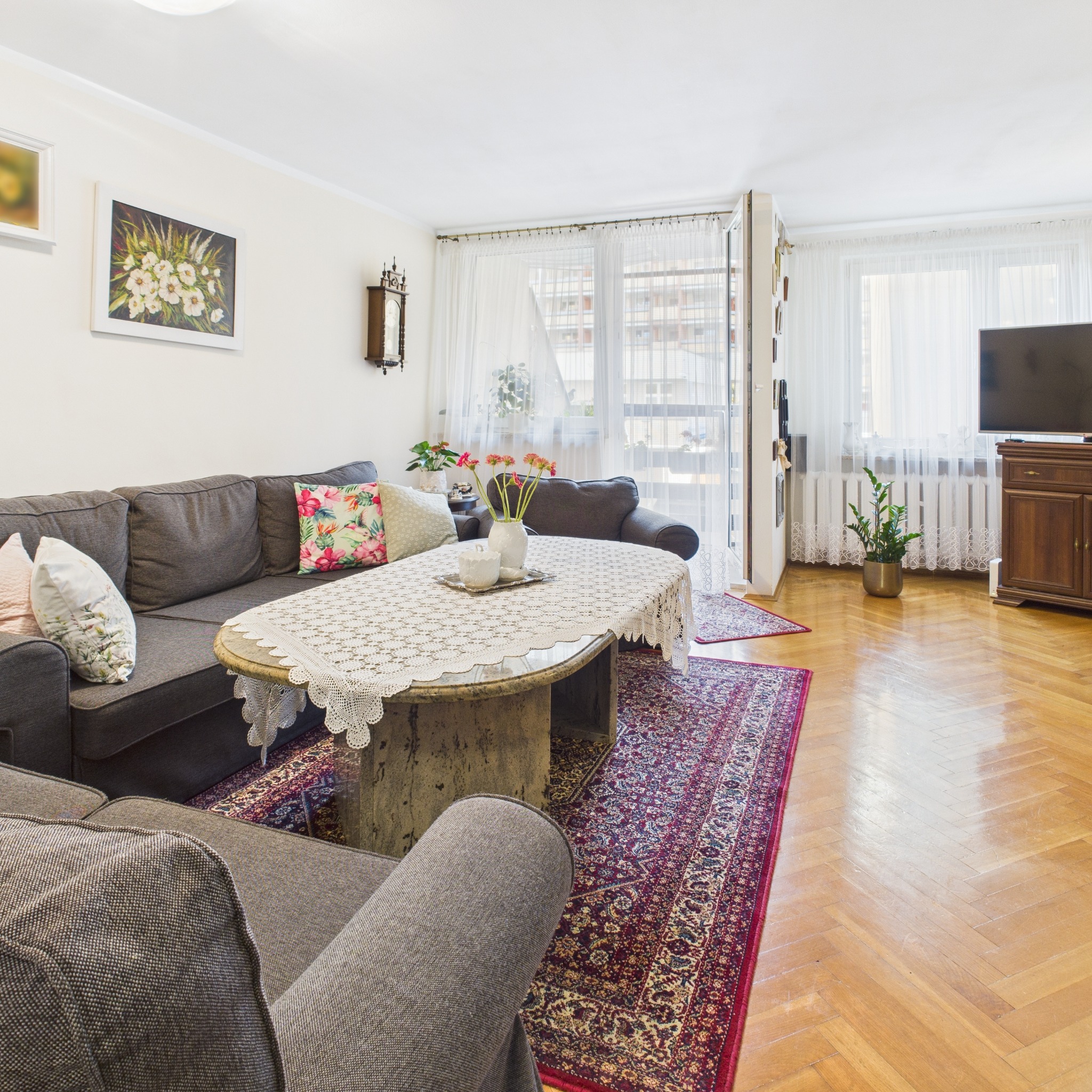 Przytulny salon z szarym narożnikiem, wzorzystym dywanem i drewnianą podłogą w jodełkę. Na ścianie obrazy i zegar, a przez okno widok na budynek mieszkalny.