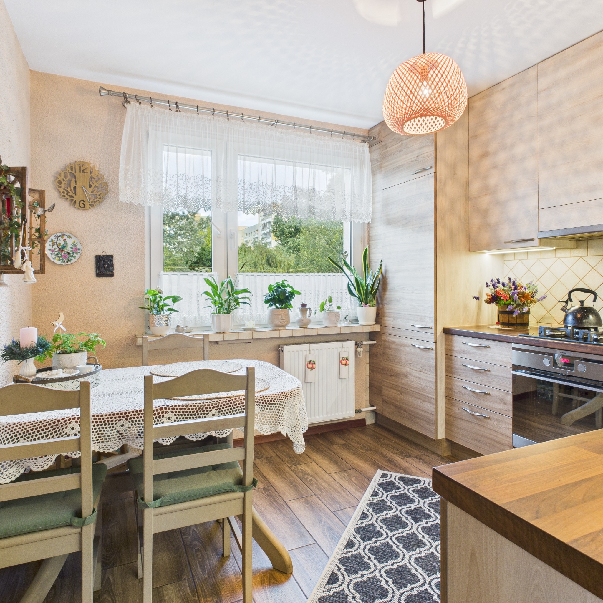 Jasna kuchnia z drewnianymi elementami, okrągłym stołem z koronkowym obrusem i roślinami na parapecie. Wnętrze doświetlone wiszącą lampą z abażurem.