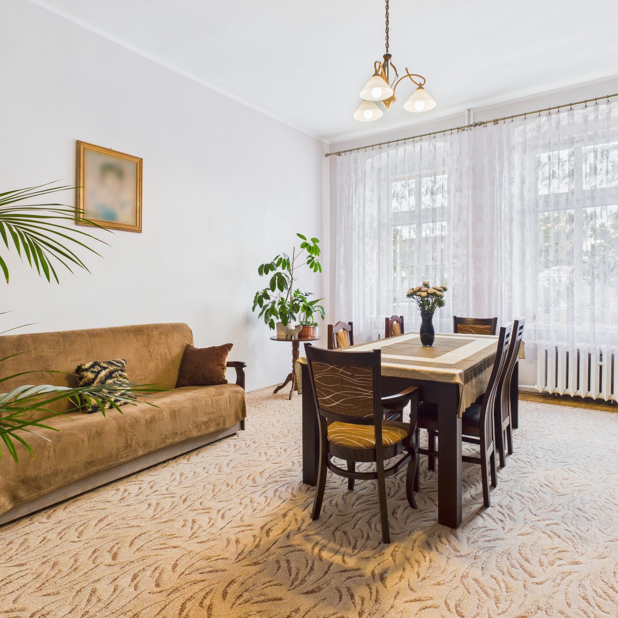 Jasne wnętrze pokoju dziennego z sofą, stołem jadalnym i kwiatami. Widoczne okno z firankami, dywan w kremowych odcieniach i dekoracje roślinne.