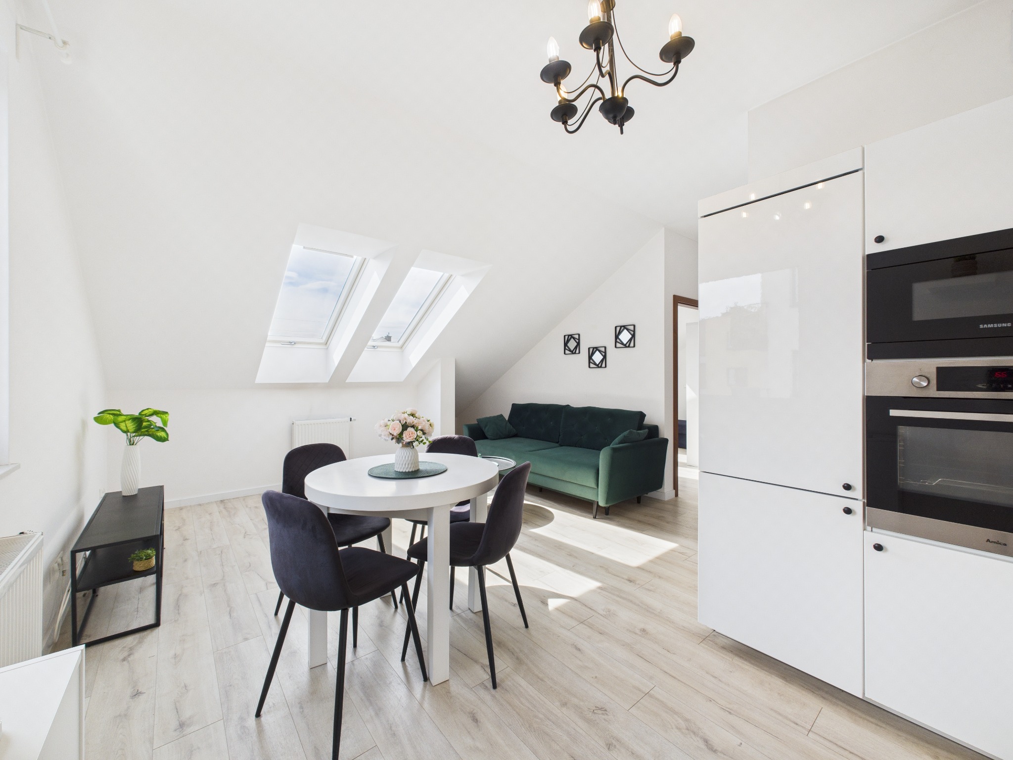 Jasne wnętrze poddasza ze stołem, krzesłami i zieloną sofą. Dwa okna dachowe wpuszczają światło. Minimalistyczna kuchnia z białymi szafkami po prawej.