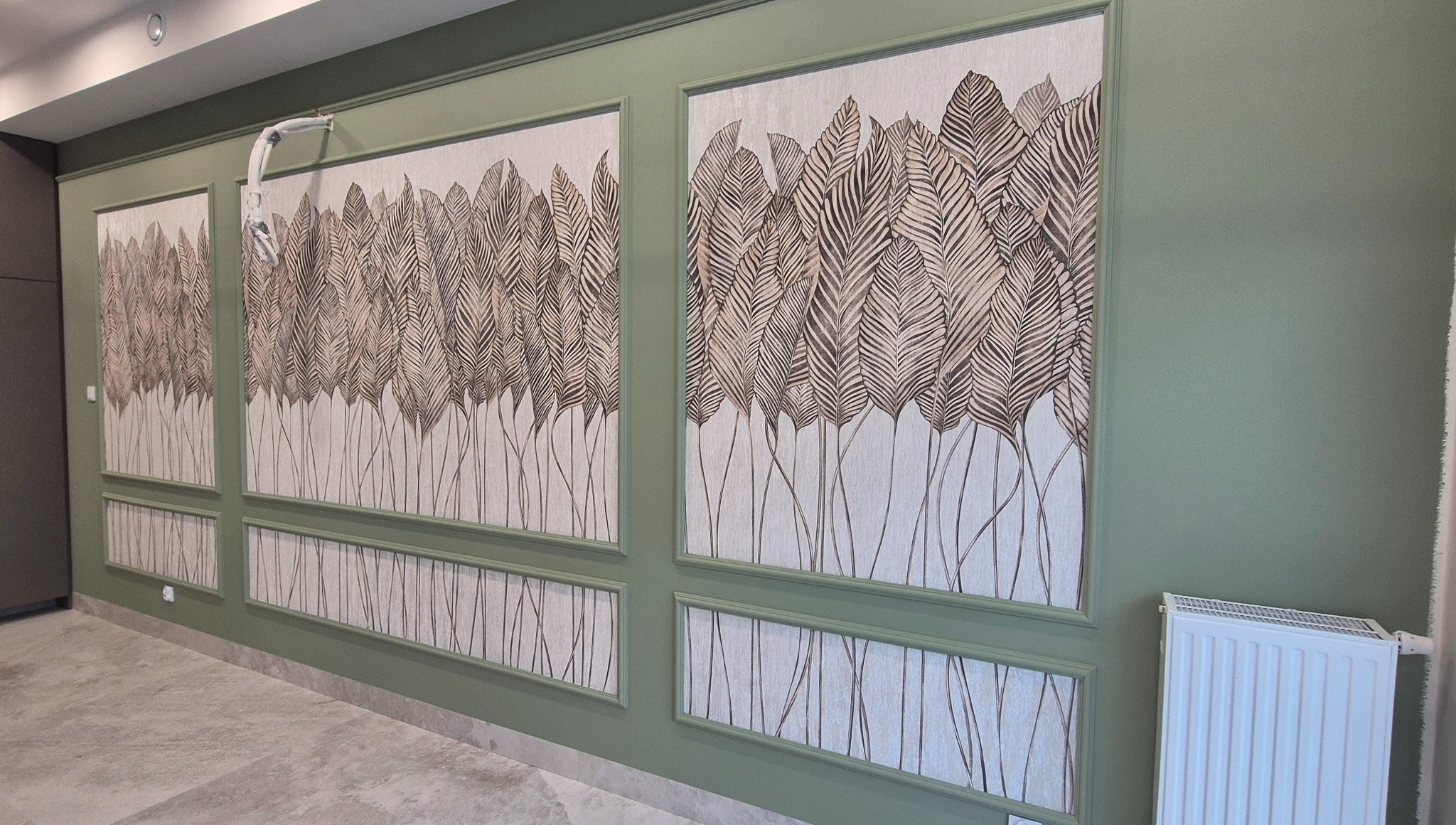 Stylowe panele ścienne z motywem liści w odcieniach brązu, umieszczone w ozdobnych ramach na ścianie w kolorze oliwkowym, z widocznym grzejnikiem.