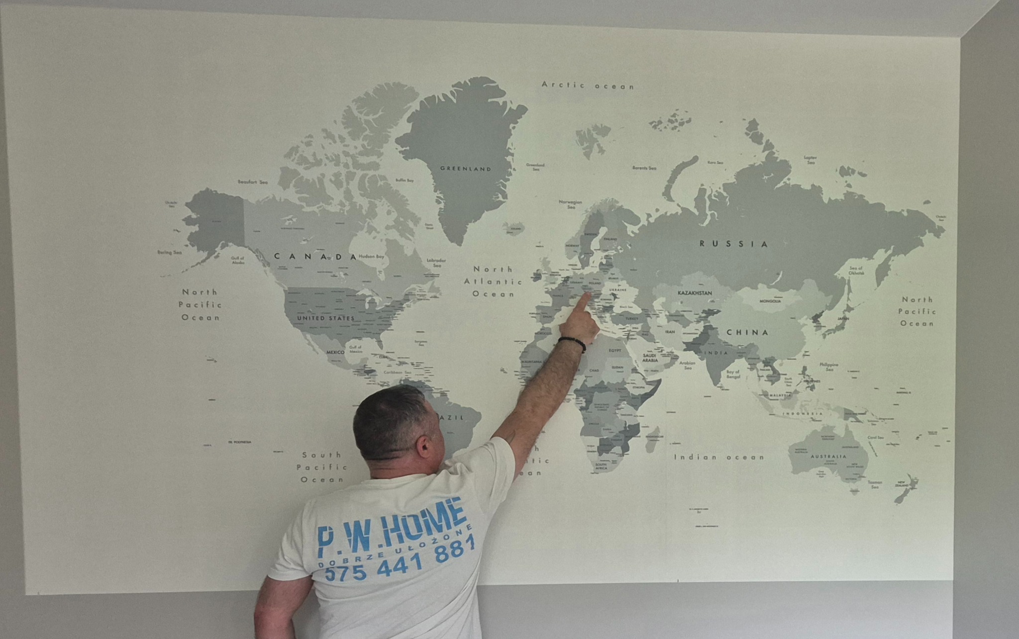 Ściana z tapetą w mapę świata, mężczyzna wskazujący palcem na Europę. Wykończenie wnętrza z unikalnym wzorem tapety, jasne tło, widoczny fragment koszulki z logo firmy.