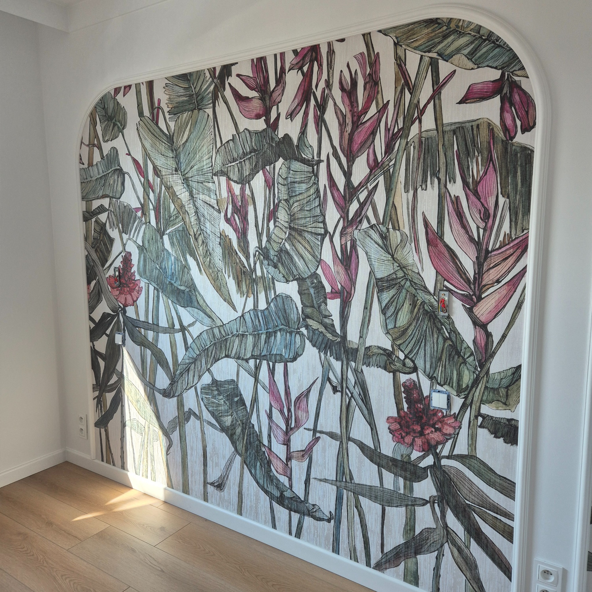 Ściana z tropikalną tapetą w łukowym wycięciu, widok na fragment podłogi z parkietem, gniazdka elektryczne po bokach, subtelne światło słoneczne.