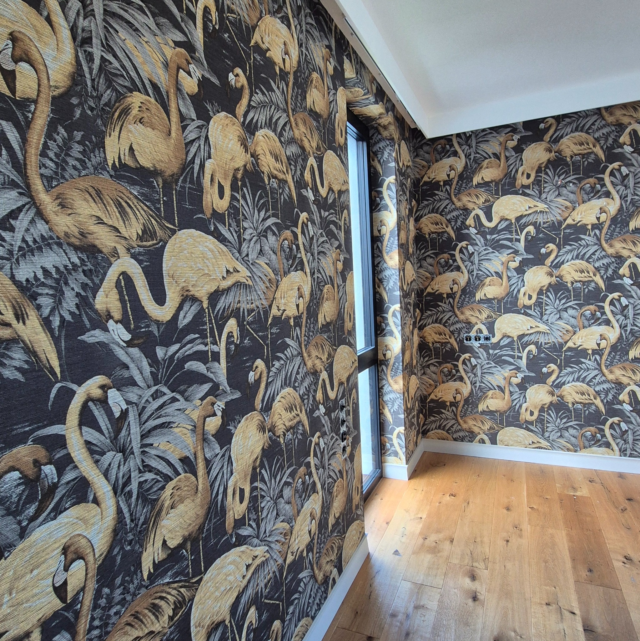 Pokój po remoncie: ściany zdobi tapeta w tropikalne flamingi na ciemnym tle, kontrastująca z jasną drewnianą podłogą i białymi listwami przypodłogowymi.