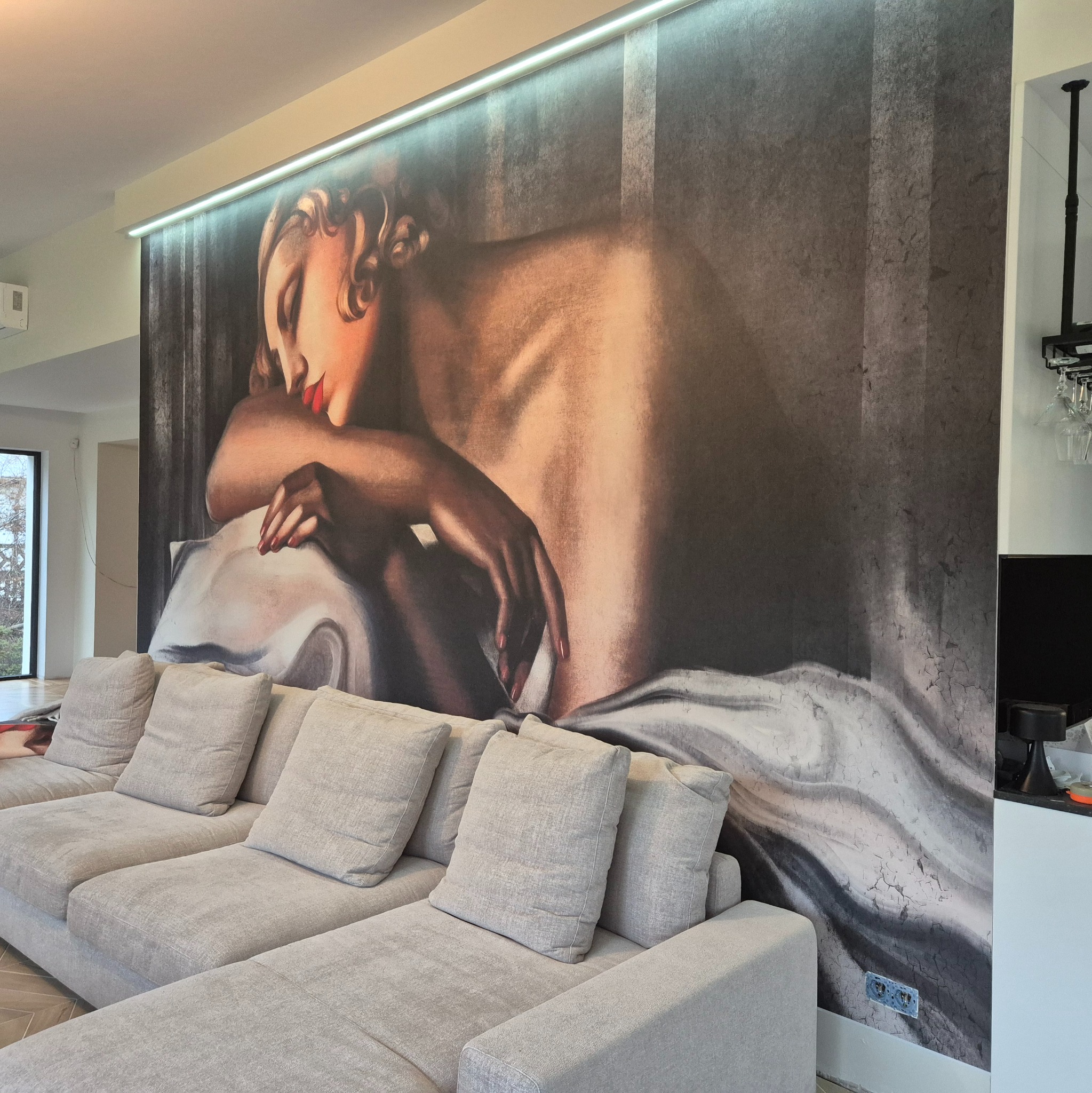 Fototapeta z wizerunkiem kobiety w secesyjnym stylu, zamontowana na ścianie w salonie nad szarą sofą. Widoczne gniazdka elektryczne w dolnej części fototapety.