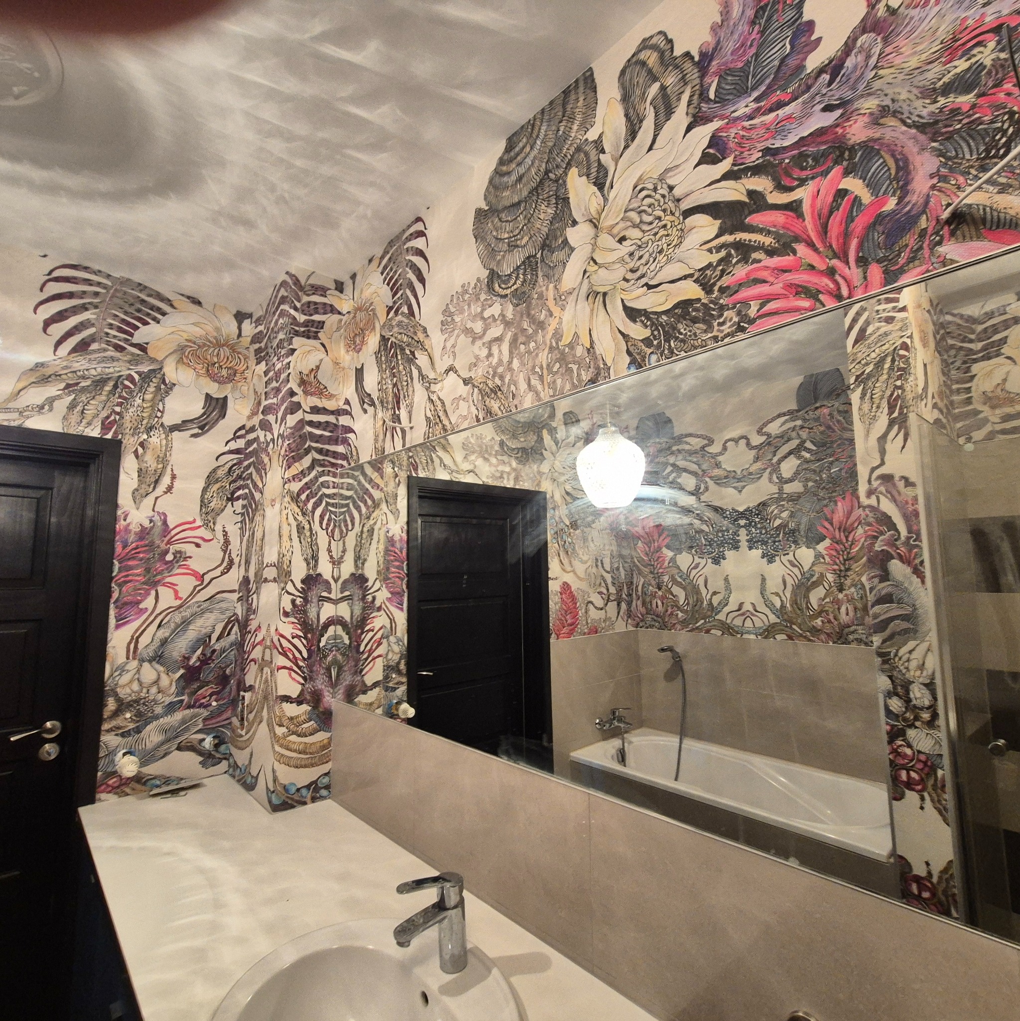 Wzorzysta tapeta w łazience z motywami kwiatowymi i roślinnymi, odbicie w lustrze. Ciemne drzwi po lewej stronie. Umywalka i bateria na pierwszym planie.