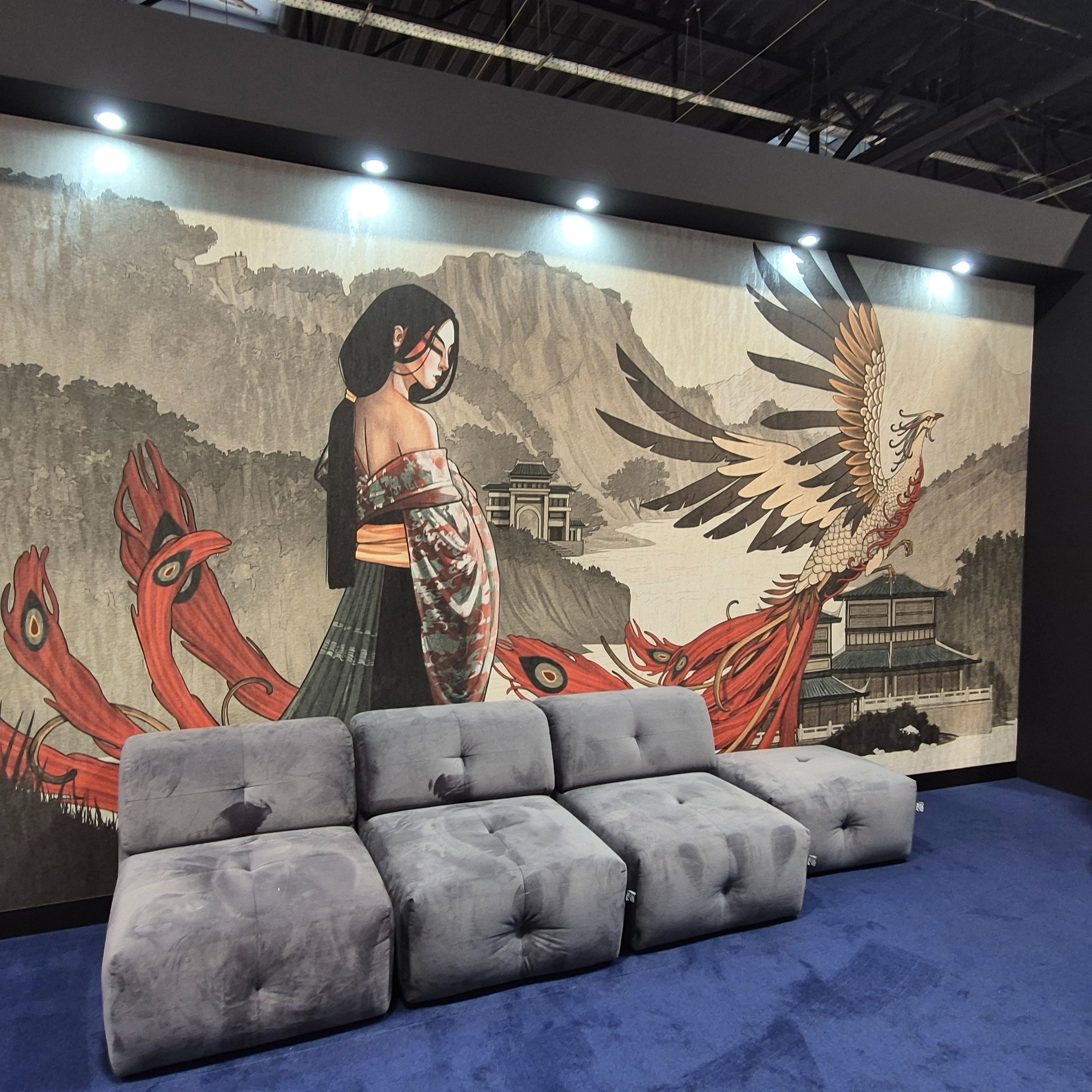 Orientalna tapeta z motywem kobiety, ptaka Feniksa i gór, oświetlona punktowo, w przestrzeni z szarą sofą i niebieską wykładziną.