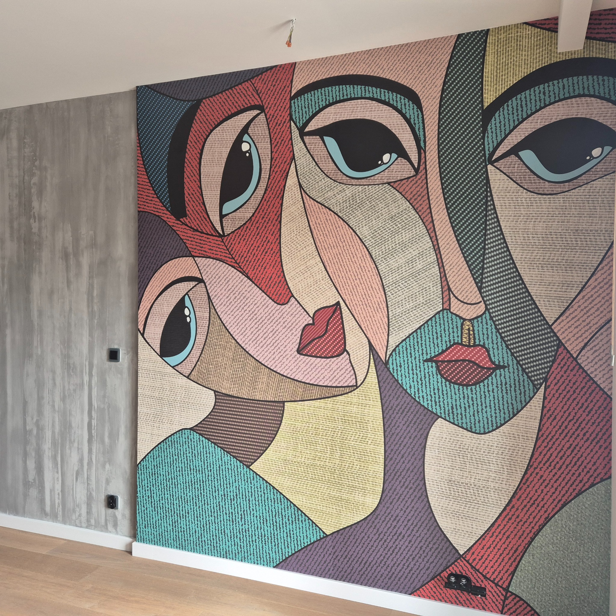 Wzorzysta tapeta z motywem abstrakcyjnych twarzy w stylu kubistycznym, nałożona na ścianę w nowoczesnym wnętrzu z szarą, betonową ścianą obok.