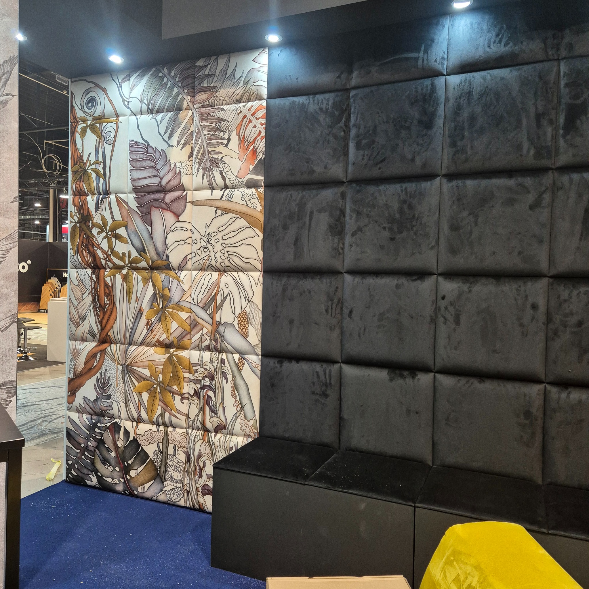 Wykończona ściana z tapicerowanymi panelami: wzór z motywami roślinnymi po lewej, ciemnoszary welur po prawej. Nowoczesny design wnętrza, oświetlenie punktowe.