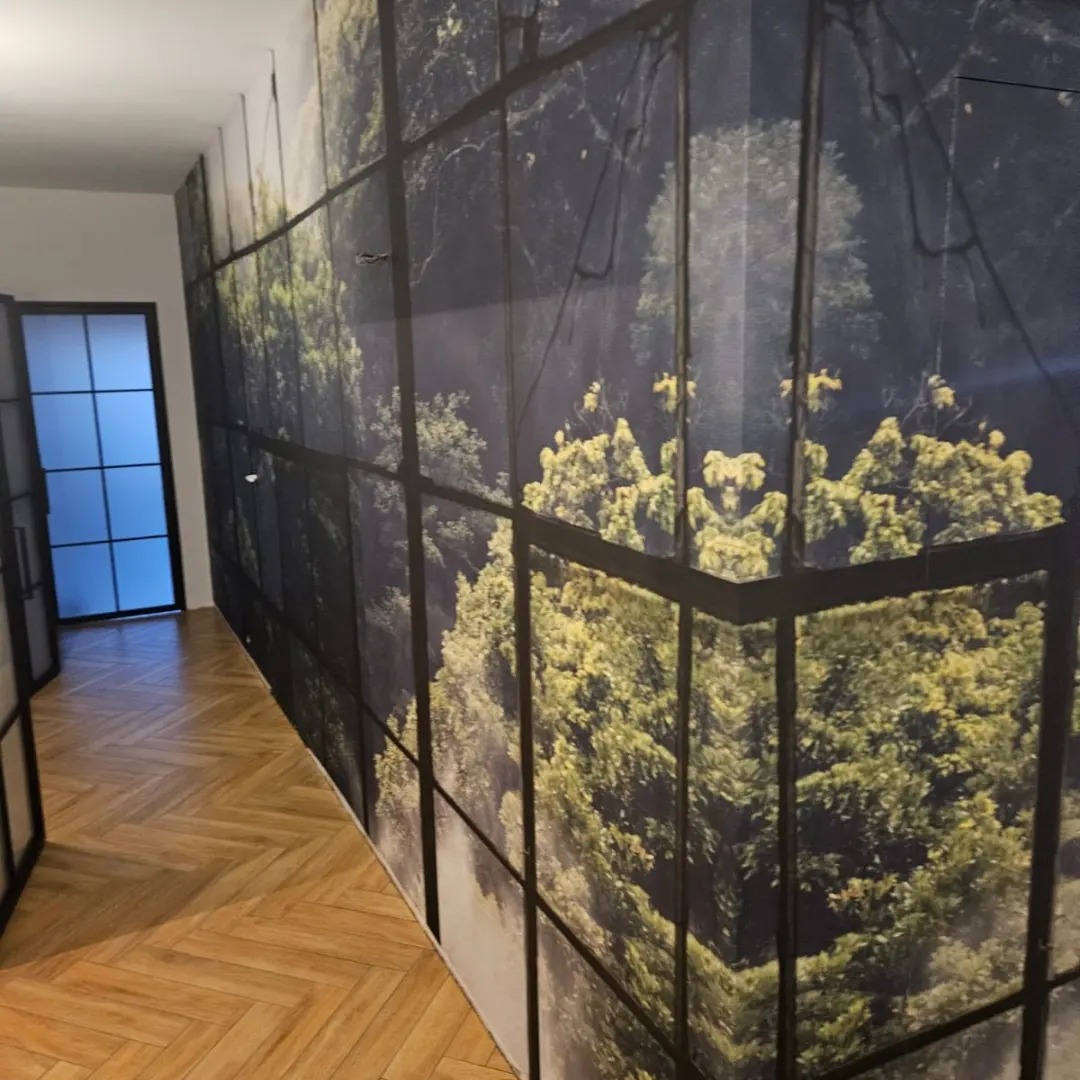 Długi korytarz z parkietem w jodełkę i fototapetą z motywem lasu za oknem, która pokrywa całą ścianę. Czarne ramy drzwi po lewej stronie.