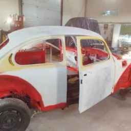 Klasyczny Volkswagen Garbus w trakcie renowacji blacharsko-lakierniczej w warsztacie. Czerwono-białe elementy karoserii, otwarte drzwi, widoczne wnętrze.