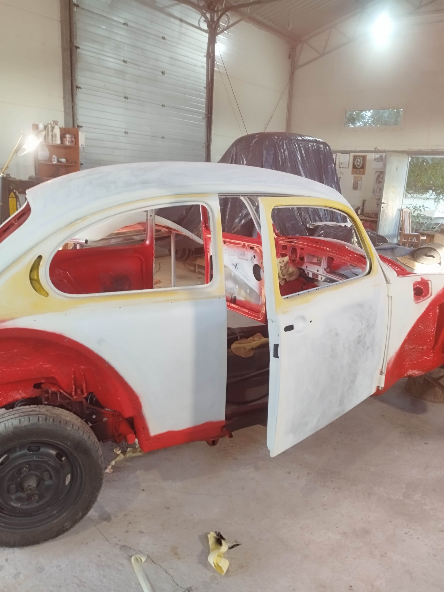 Klasyczny Volkswagen Garbus w trakcie renowacji blacharsko-lakierniczej w warsztacie. Czerwono-białe elementy karoserii, otwarte drzwi, widoczne wnętrze.