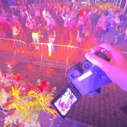HuBrt - Kamerzysta filmuje bawiący się tłum pod namiotem, widok z ręki trzymającej kamerę, w tle kwiaty i barierki. Scena oświetlona fioletowym światłem.