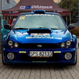 HuBrt - Błękitny Subaru World Rally Team z numerem 126 na tle namiotu Darma. Widoczne logo Motul i RR Customs. Zdjęcie wykonane z przodu, na brukowanej nawierzchni.