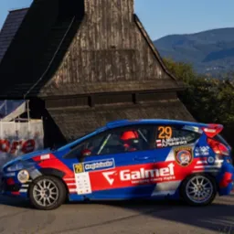 Niebieski rajdowy Ford Fiesta z numerem 29 na tle drewnianego kościoła i gór. Widoczny kierowca w kasku. Galmet i #rajdslaska na karoserii.