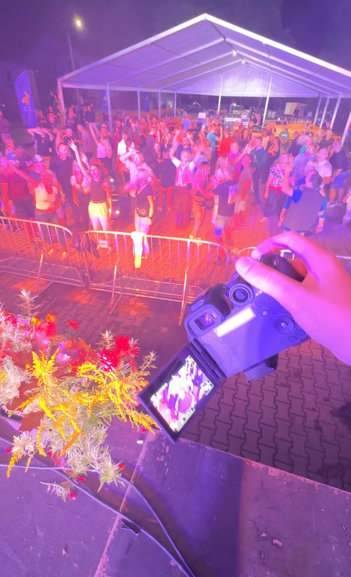 Kamerzysta filmuje bawiący się tłum pod namiotem, widok z ręki trzymającej kamerę, w tle kwiaty i barierki. Scena oświetlona fioletowym światłem.