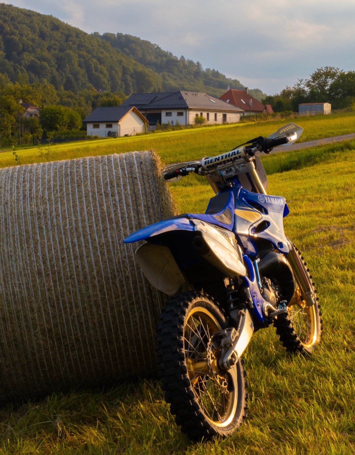 Niebieski motocykl Yamaha oparty o belę siana na łące, w tle domy i zalesione wzgórze oświetlone zachodzącym słońcem. Ujęcie z dołu podkreśla wielkość motocykla.