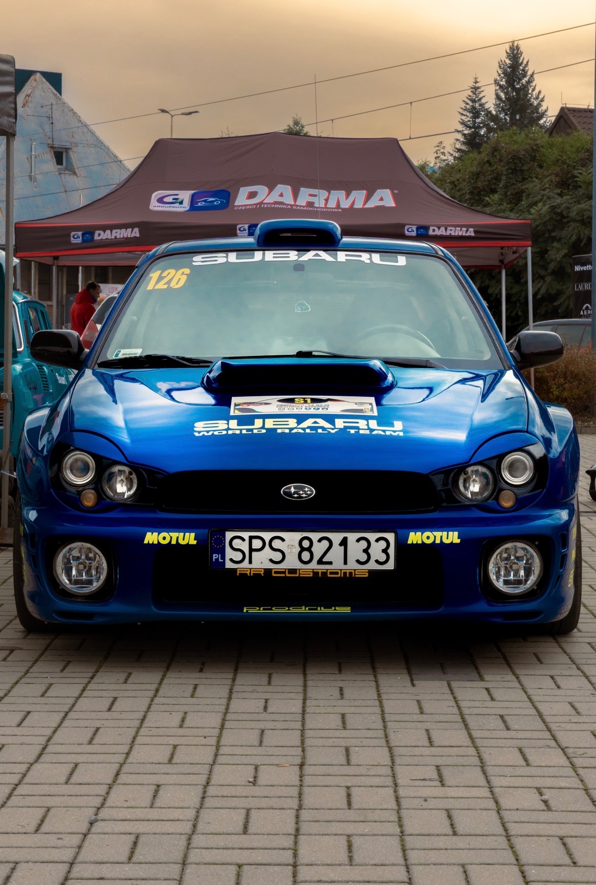 Błękitny Subaru World Rally Team z numerem 126 na tle namiotu Darma. Widoczne logo Motul i RR Customs. Zdjęcie wykonane z przodu, na brukowanej nawierzchni.