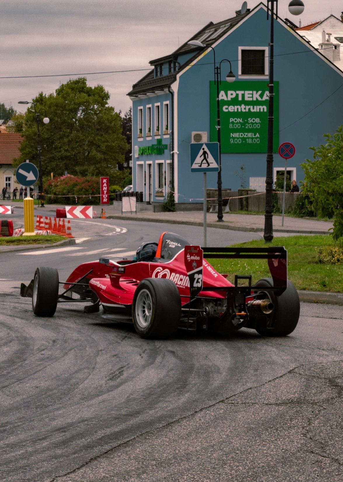 Czerwony bolid wyścigowy na tle miejskiego krajobrazu z budynkiem apteki. Ujęcie z tyłu, widoczne detale konstrukcji i numer 23. Asfaltowa droga.