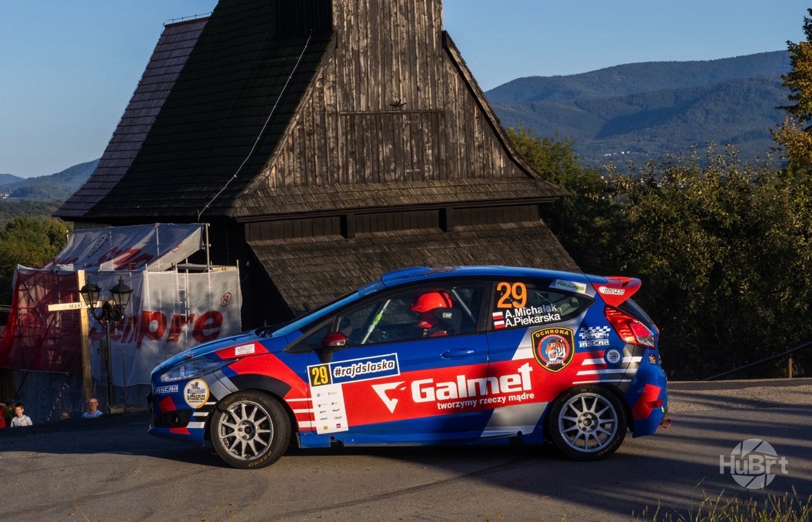 Niebieski rajdowy Ford Fiesta z numerem 29 na tle drewnianego kościoła i gór. Widoczny kierowca w kasku. Galmet i #rajdslaska na karoserii.