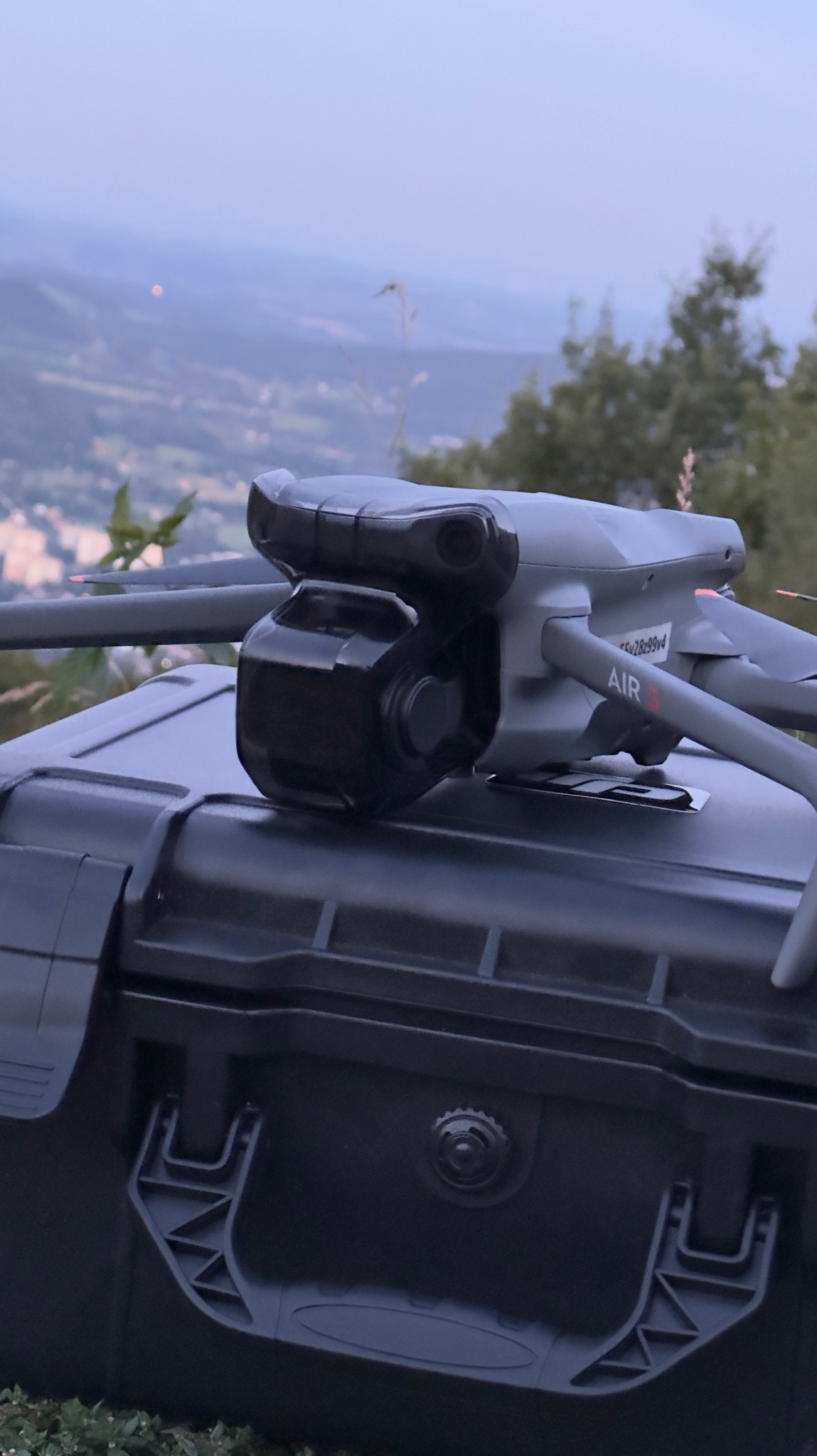 Szary dron DJI Air 3 spoczywa na czarnym, twardym futerale na tle rozmytego krajobrazu górskiego o zmierzchu. Widoczny numer seryjny drona.