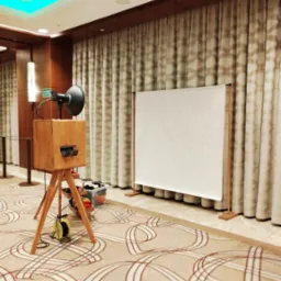 Drewniana fotobudka z lampą i rekwizytami stoi na tle kremowej zasłony i wzorzystego dywanu w eleganckim wnętrzu, gotowa do użycia podczas imprezy.