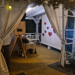 MSM GRUPA MACIEJ SKUPIEWSKI - Drewniana fotobudka z dekoracjami (serca, zasłony) na zewnątrz, oświetlona girlandami żarówek, ustawiona na tarasie z kostki brukowej podczas imprezy.