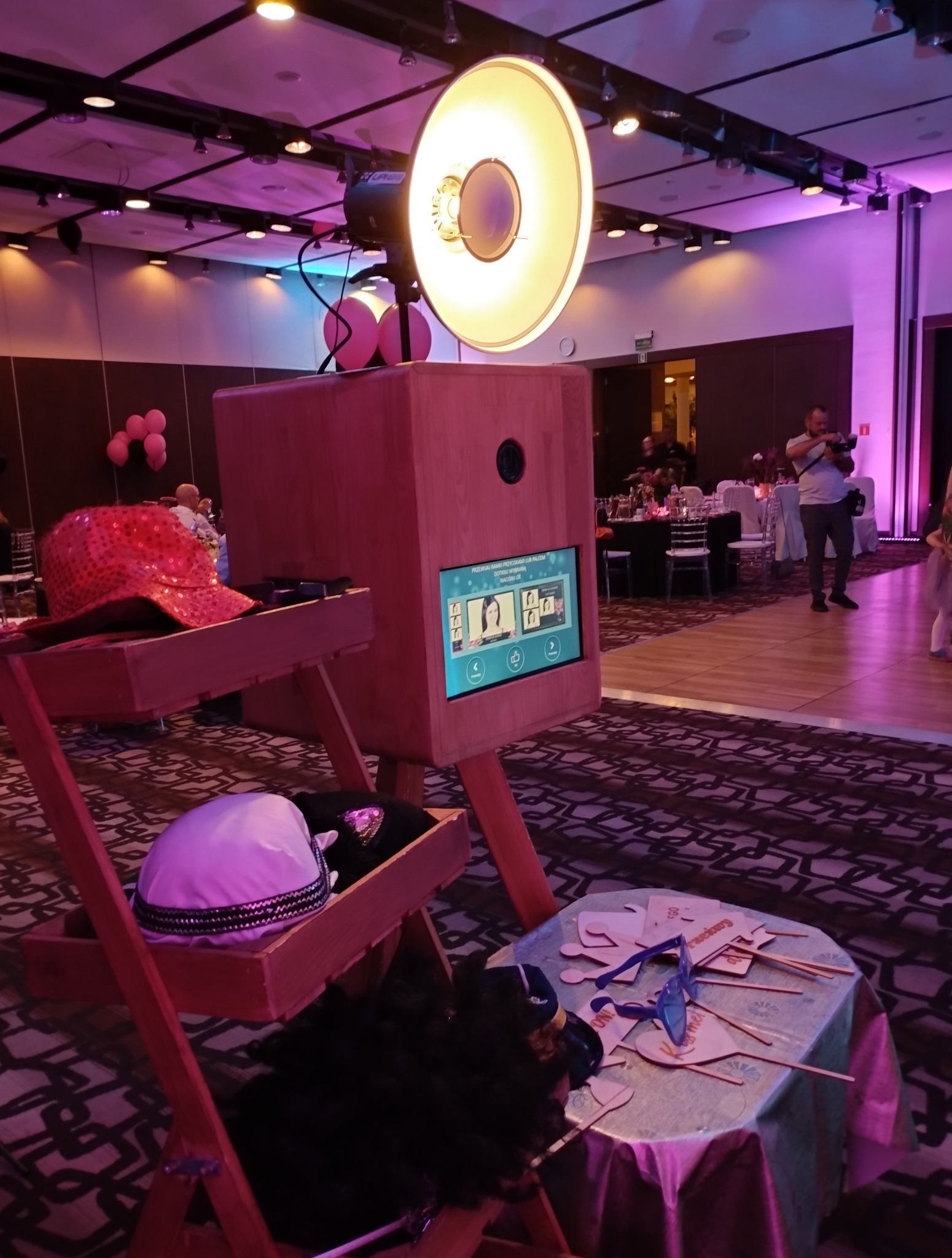 Drewniana fotobudka z ekranem dotykowym, rekwizytami (kapelusze, okulary) na stole. W tle sala bankietowa oświetlona na różowo.