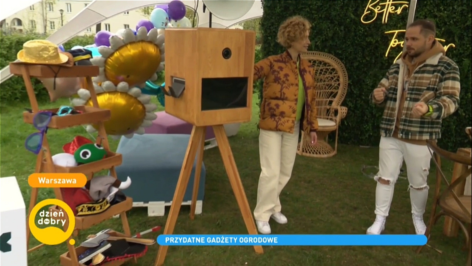 Plenerowa fotobudka z rekwizytami, otoczona osobami i dekoracjami, na tle zieleni. Drewniana konstrukcja na trójnogu, rekwizyty na wózku, neon 'Better Together'.