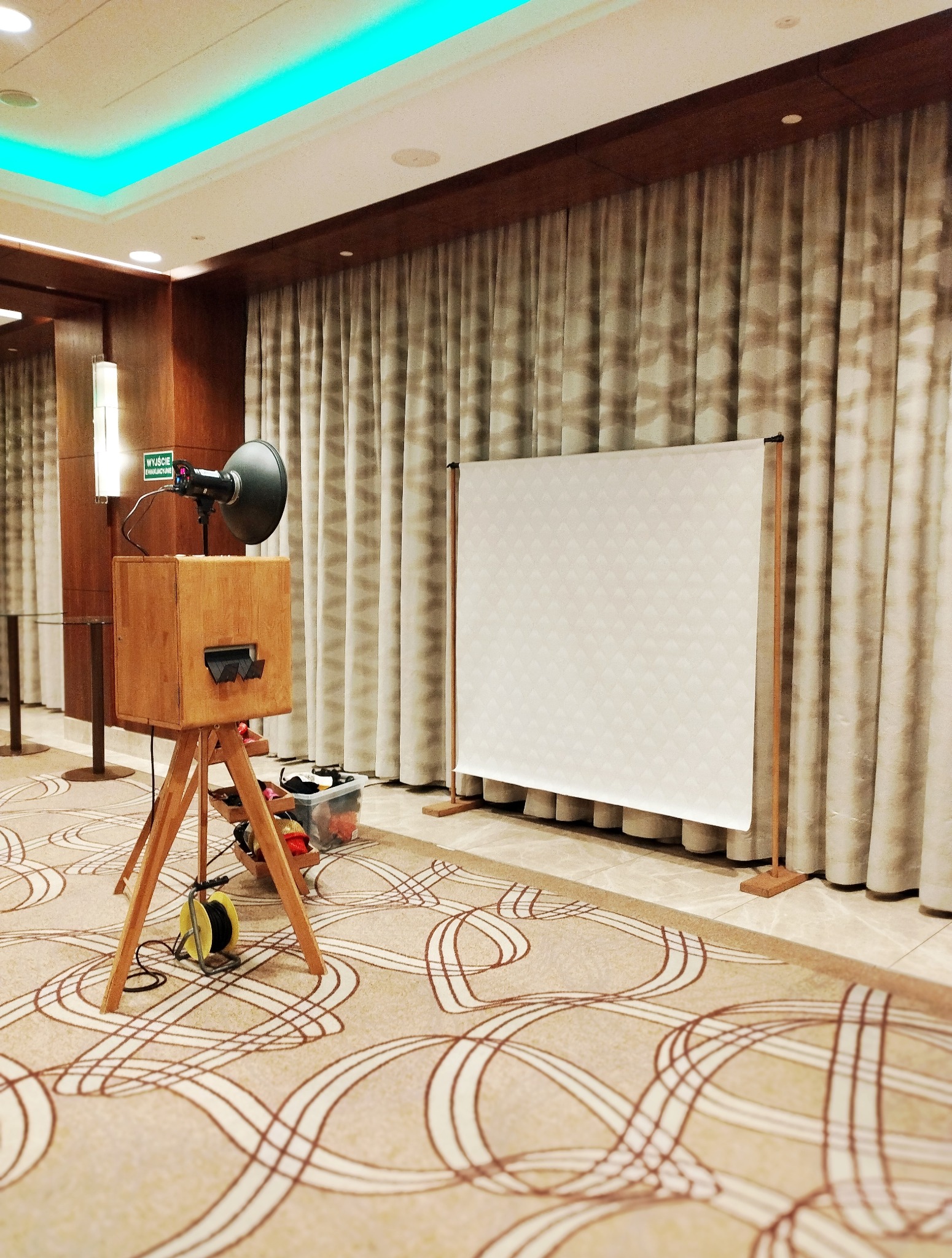 Drewniana fotobudka z lampą i rekwizytami stoi na tle kremowej zasłony i wzorzystego dywanu w eleganckim wnętrzu, gotowa do użycia podczas imprezy.