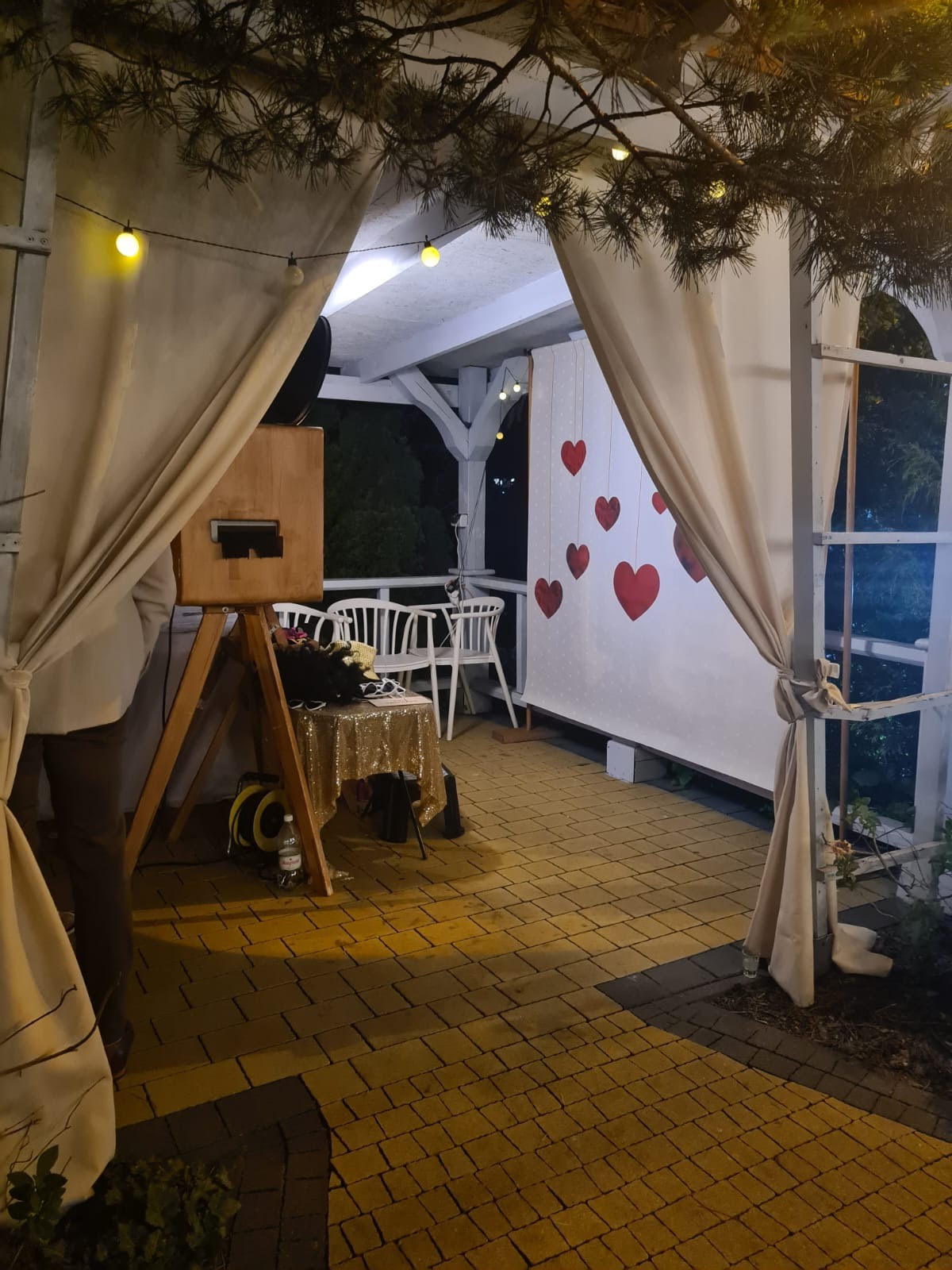 Drewniana fotobudka z dekoracjami (serca, zasłony) na zewnątrz, oświetlona girlandami żarówek, ustawiona na tarasie z kostki brukowej podczas imprezy.
