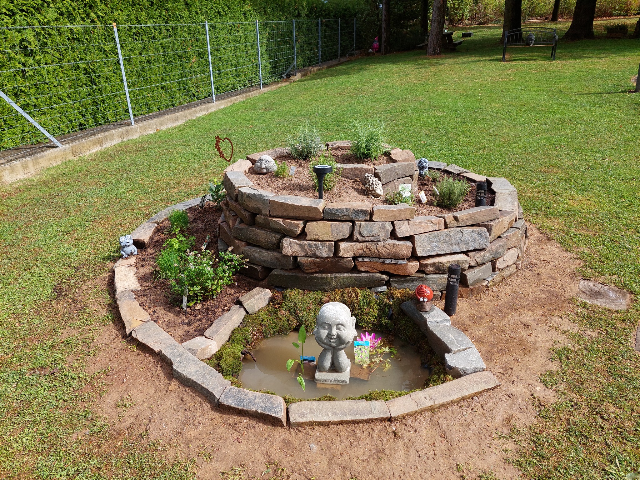 Ogród z oczkiem wodnym i kamienną obudową, dekorowany figurkami i roślinami. W tle trawnik i żywopłot, tworzące spójną kompozycję.