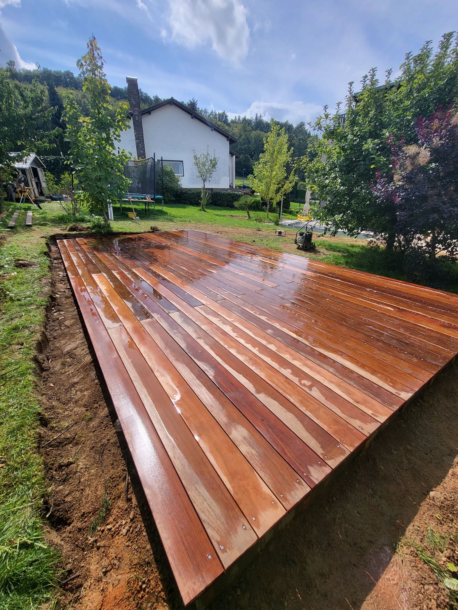 Świeżo ułożony, mokry drewniany taras w ogrodzie, z widocznymi śladami ziemi po obrysie, w tle dom i zieleń. Deski ułożone równolegle, widoczne śruby mocujące.