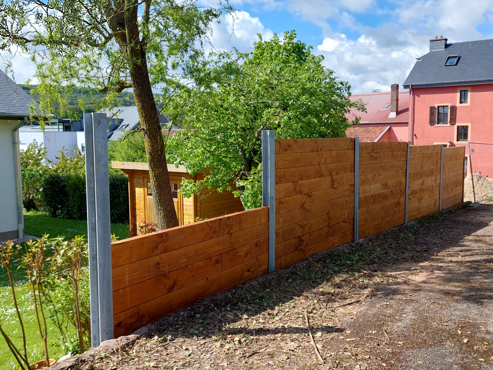 Nowy, drewniany płot z metalowymi słupkami oddziela posesję. W tle widoczny ogród, drzewa i budynki mieszkalne w słoneczny dzień.