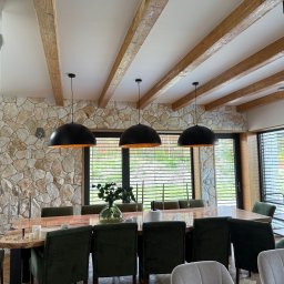 M&M MiraMark Vitalii Deputatov - Jadalnia z kamienną ścianą, drewnianymi belkami i czarnymi lampami. Stół z ciemnego drewna otoczony zielonymi krzesłami. Widoczne okna z żaluzjami.