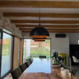 Stylowa jadalnia z drewnianym stołem o nieregularnym kształcie i zielonymi krzesłami. Widoczne drewniane belki stropowe i kamienna ściana. Okno z żaluzjami w tle.