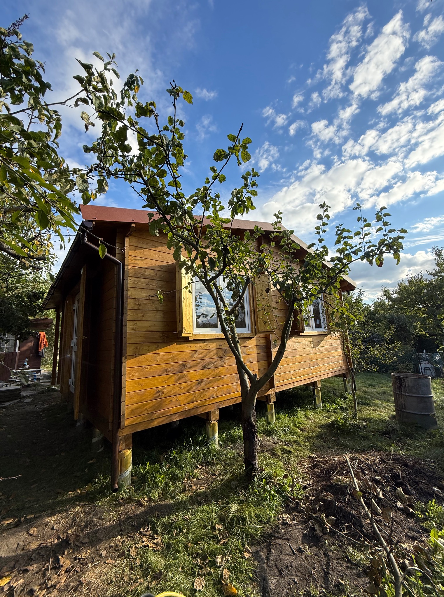 Drewniany domek letniskowy z białymi oknami, na podwyższeniu, otoczony zielenią, pod błękitnym niebem z chmurami. Widok z boku, w naturalnym oświetleniu.
