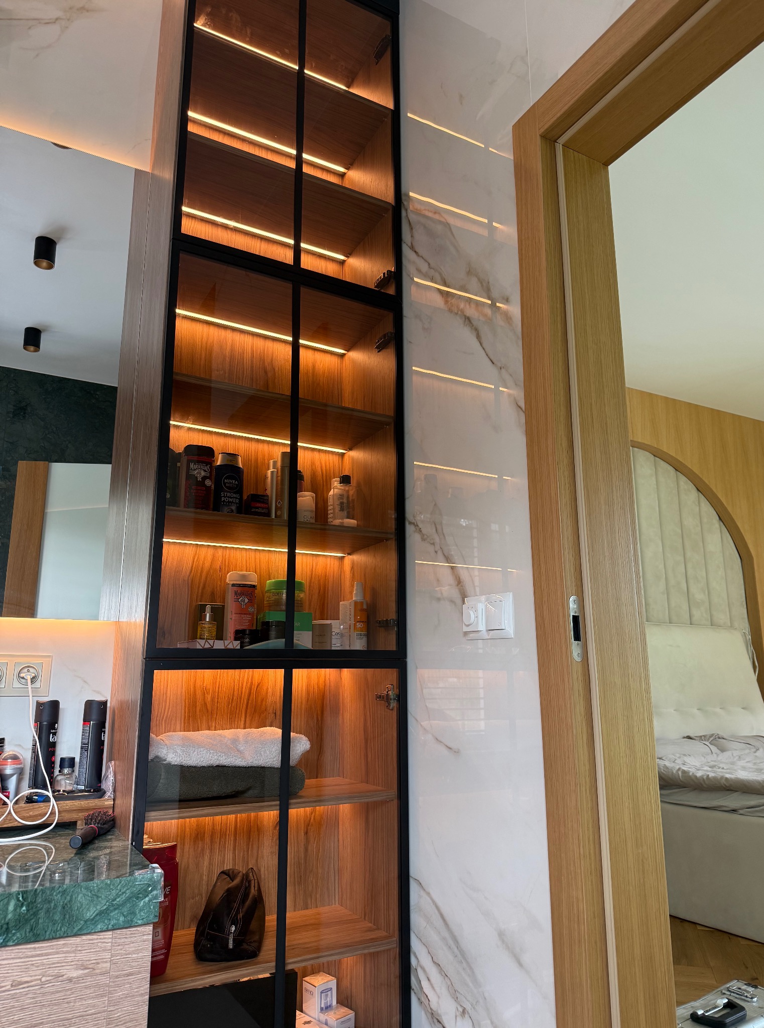 Szklana witryna z oświetleniem LED w łazience, z kosmetykami i ręcznikami. Marmurowe wykończenie ściany i fragment drzwi. Widok na sypialnię w tle.
