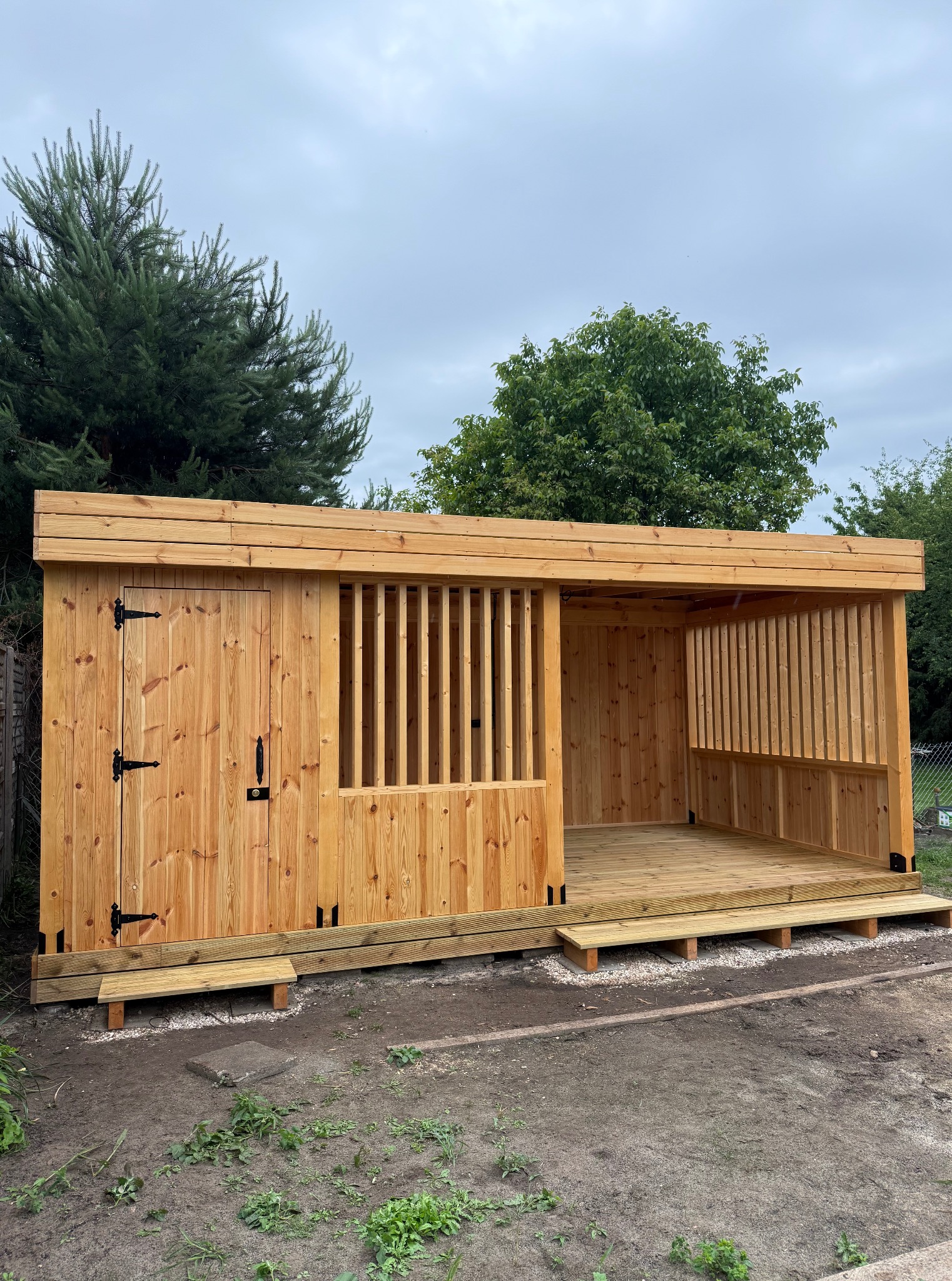 Nowoczesna, drewniana altana z zamykanym pomieszczeniem i ażurową ścianą, świeżo po montażu, na tle zieleni. Widoczne detale wykończenia i naturalna kolorystyka drewna.