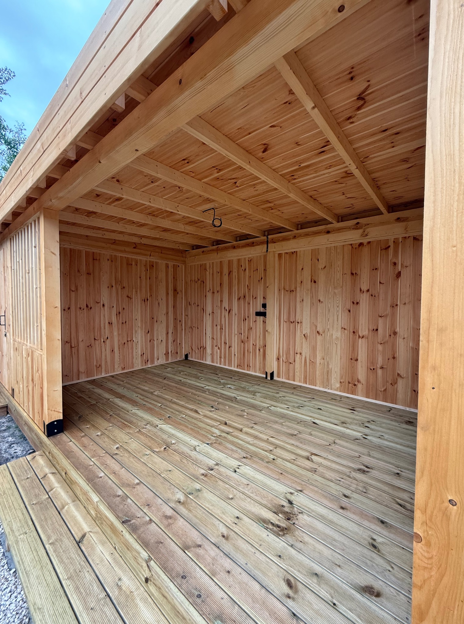 Drewniana konstrukcja altany z desek, widok z zewnątrz. Ściany, podłoga i sufit wykonane z jasnego drewna. Widoczne detale łączeń i naturalny rysunek drewna.