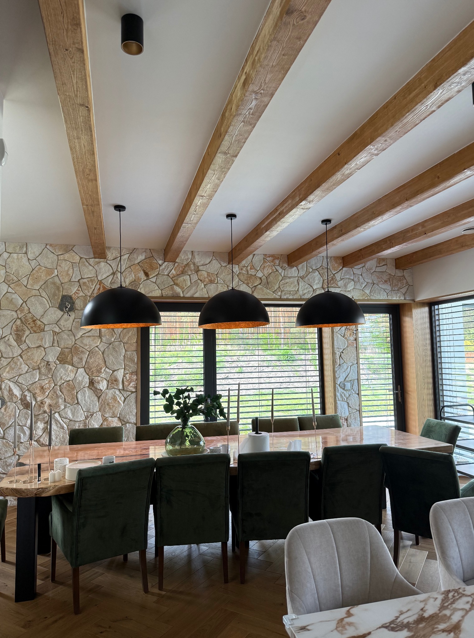 Jadalnia z kamienną ścianą, drewnianymi belkami i czarnymi lampami. Stół z ciemnego drewna otoczony zielonymi krzesłami. Widoczne okna z żaluzjami.