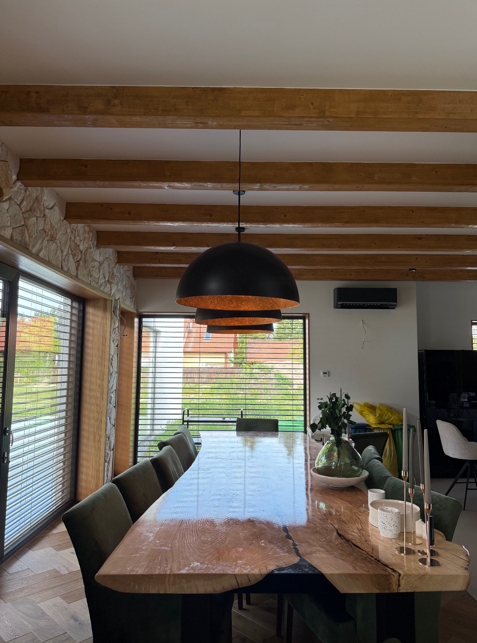 Stylowa jadalnia z drewnianym stołem o nieregularnym kształcie i zielonymi krzesłami. Widoczne drewniane belki stropowe i kamienna ściana. Okno z żaluzjami w tle.