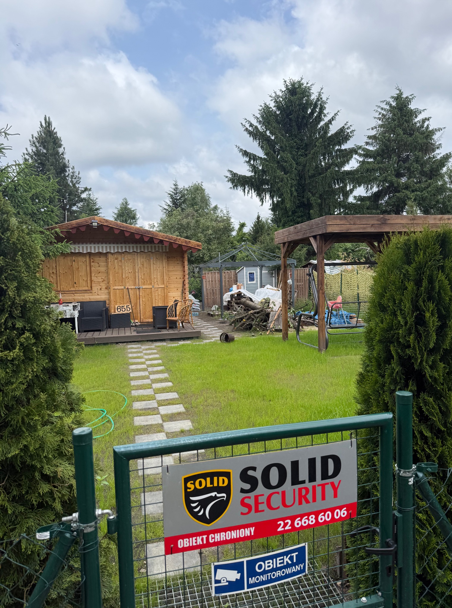 Ogród z drewnianą altaną, ścieżką z płyt i elementami małej architektury. Na pierwszym planie brama z tabliczką firmy ochroniarskiej Solid Security.