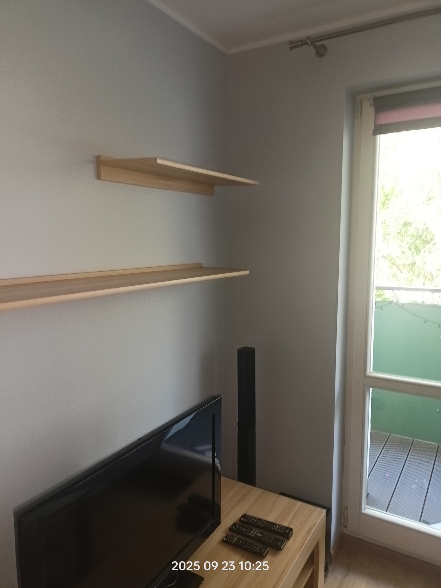 Jasne, drewniane półki na szarej ścianie nad telewizorem. Widok na balkon przez okno. Minimalistyczny wystrój wnętrza.