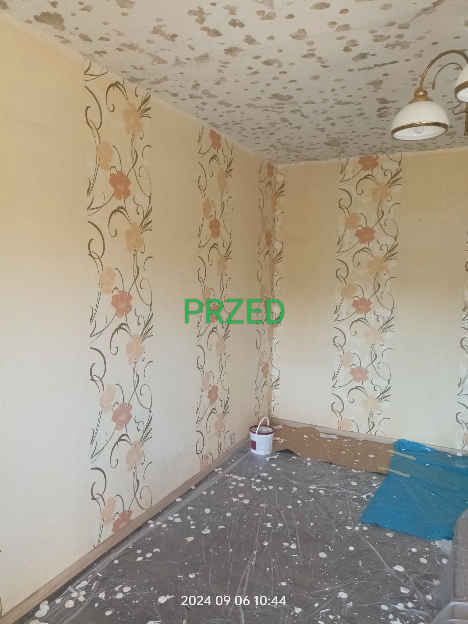 Pokój przed remontem: ściany w kwiatowe pasy, plamy na suficie, podłoga zabezpieczona folią, widoczny napis 'PRZED' i wiadro z farbą. Ujęcie z narożnika.