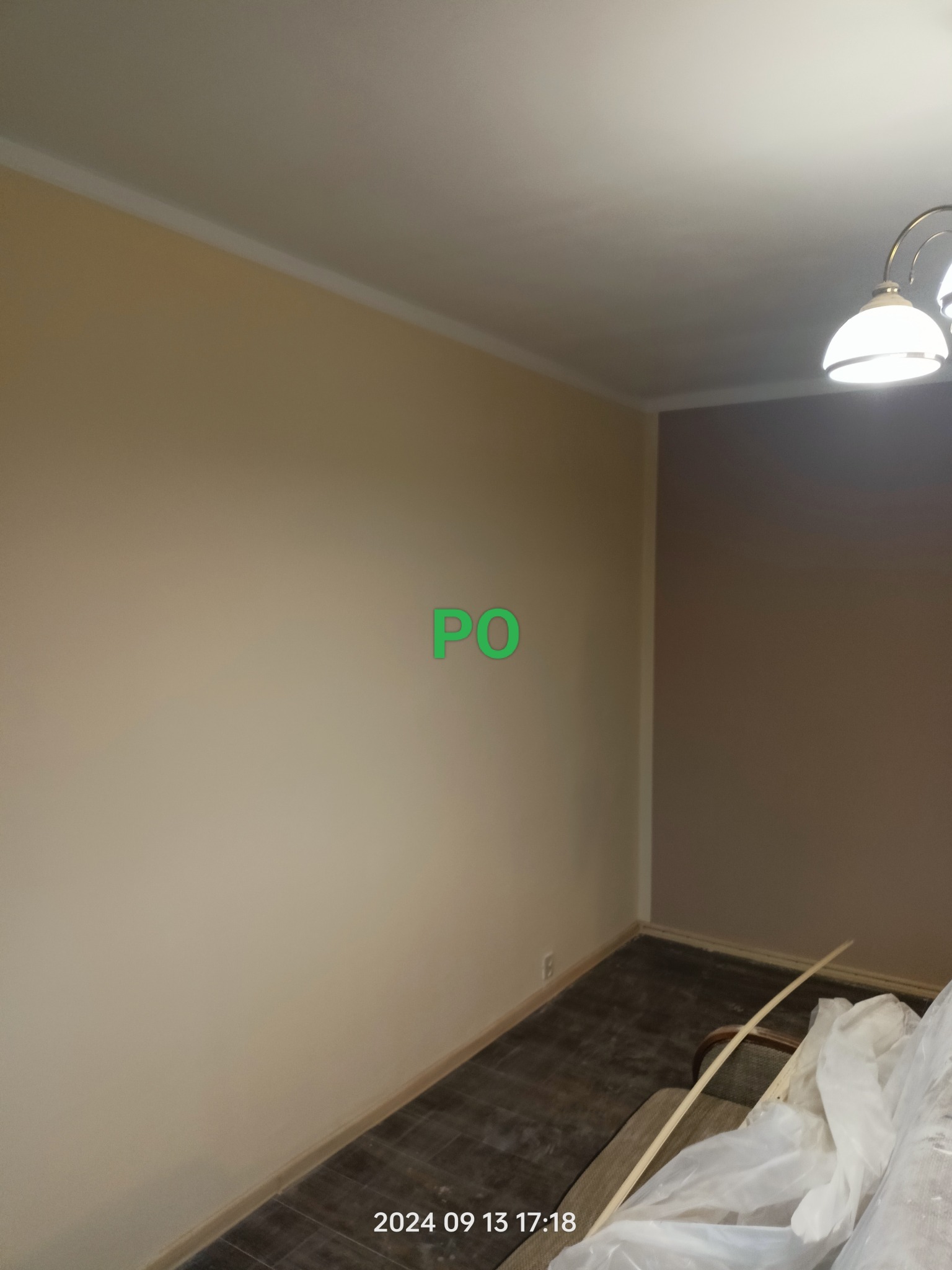 Ściana po malowaniu w dwóch odcieniach beżu, wykończona białą listwą. Częściowo widoczne materiały budowlane i lampa sufitowa. Widoczne oznaczenie 'PO'.