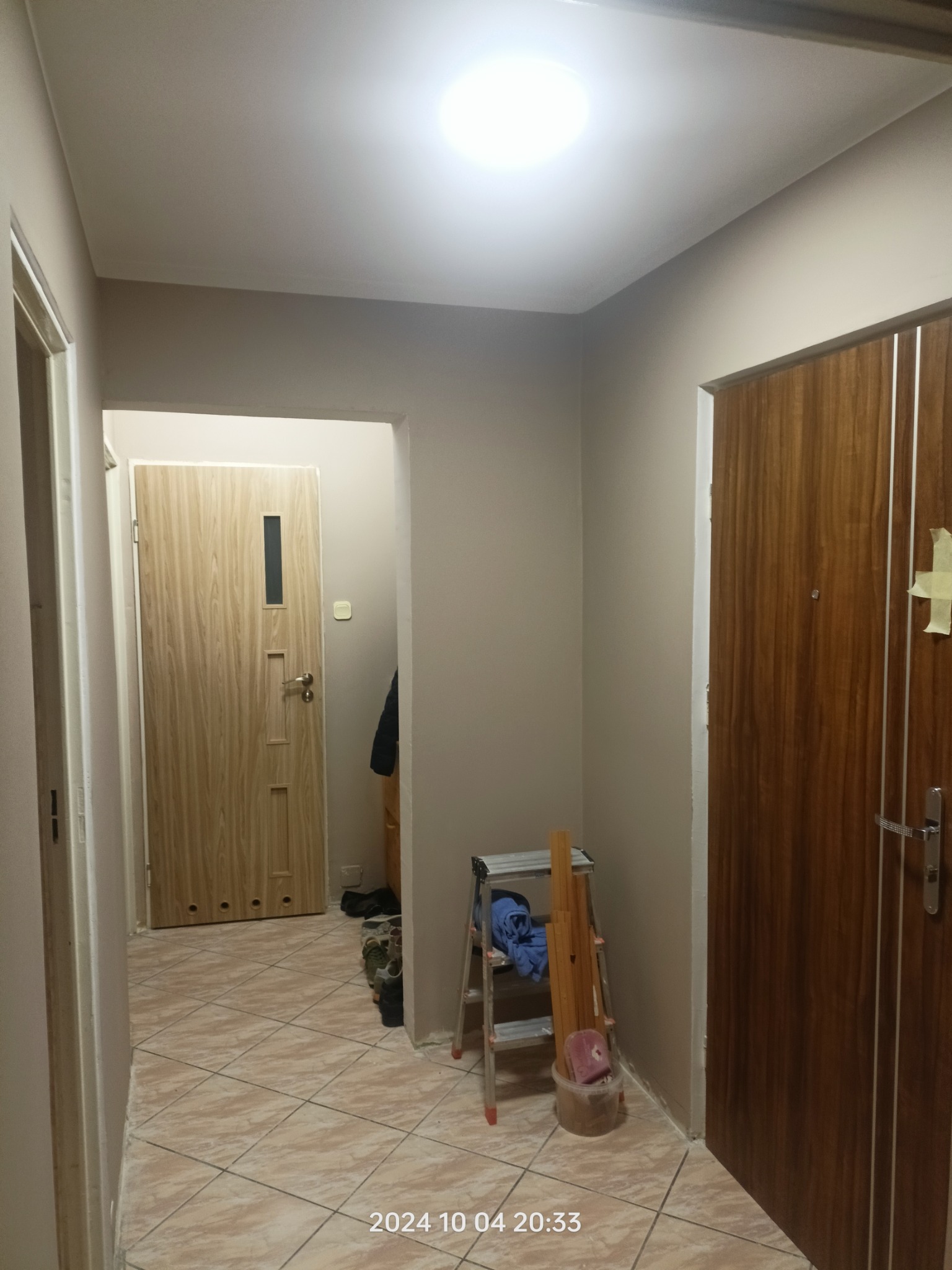 Korytarz po malowaniu na beżowo z widoczną drabiną, podłogą z płytek i dwiema parami drzwi. Na jednej z nich widoczna taśma malarska.